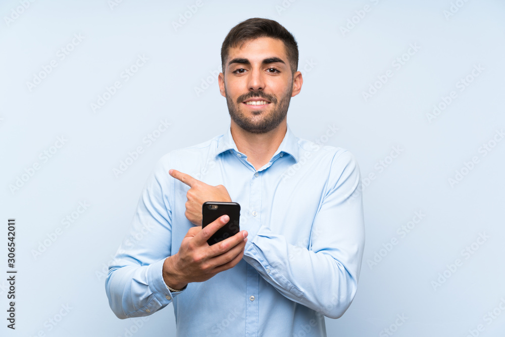 Young handsome man with his mobile over isolated blue wall pointing to the side to present a product Young handsome man with his mobile over isolated blue wall pointing to the side to present a product