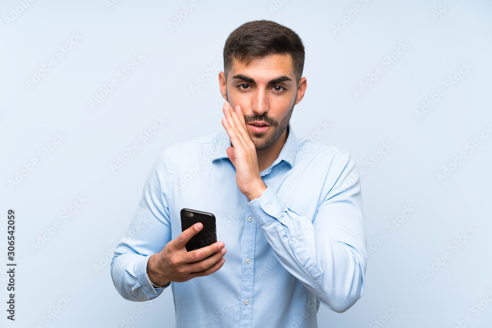 Young handsome man with his mobile over isolated blue wall whispering something Young handsome man with his mobile over isolated blue wall whispering something