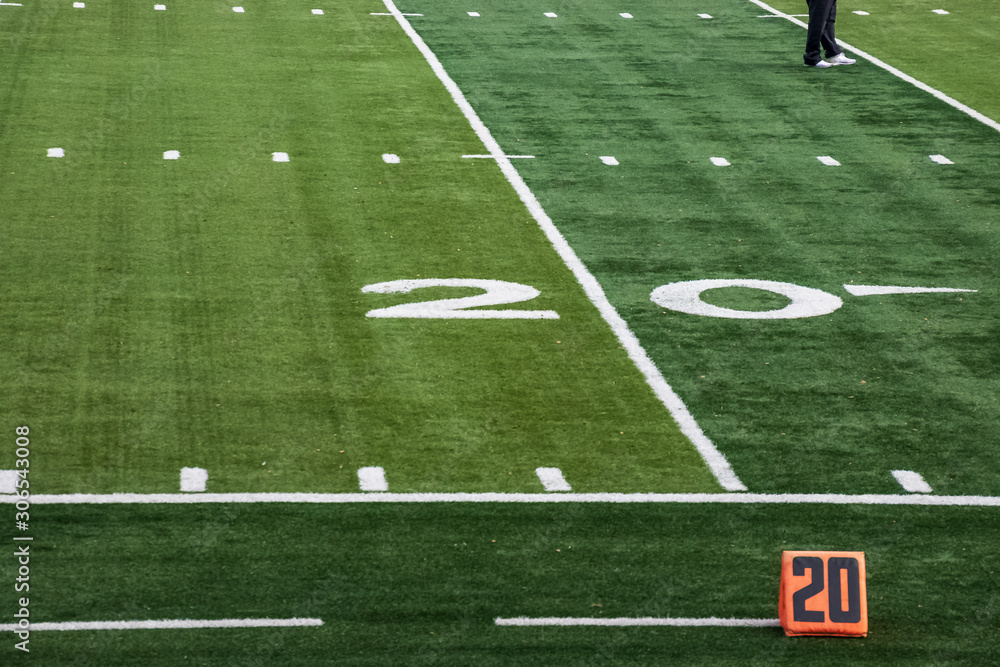 Fototapeta premium View of 20 yard line markers for American football field with feet of person standing on artificial turf