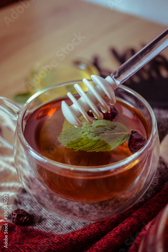 Hand holding silic honey dipper with dipping honey into glass tea cup with dried rosehip and fresh leaves mint on wooden table in color background, herbal tea and healthy drink concept. Close up. photo