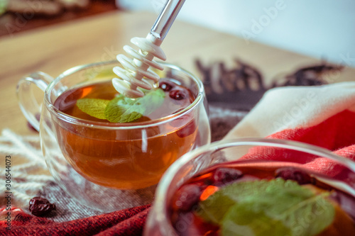 Hand holding silic honey dipper with dipping honey into glass tea cup with dried rosehip and fresh leaves mint on wooden table in color background, herbal tea and healthy drink concept. Close up. photo