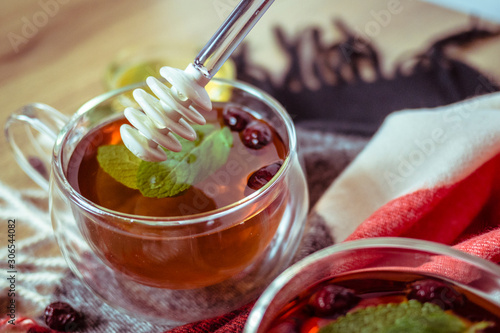 Hand holding silic honey dipper with dipping honey into glass tea cup with dried rosehip and fresh leaves mint on wooden table in color background, herbal tea and healthy drink concept. Close up. photo