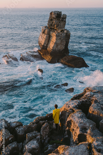Fotografie Traveler in the yellow jacket standing on the rocks in front of a huge waves