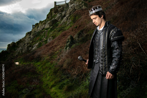 Obraz na plátně Handsome king with sword stands in contemplation with hill and parts of keep in