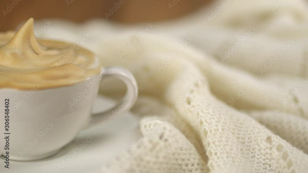 Dolly shot of a tasty cup of coffee cappuccino with a sweet cream on it on a wooden table of home or a coffee house near a white warm soft woolen blanket. Drink, beverage. Breakfast. Dolly shot of a tasty cup of coffee cappuccino with a sweet cream on it on a wooden table of home or a coffee house near a white warm soft woolen blanket. Drink, beverage. Breakfast.