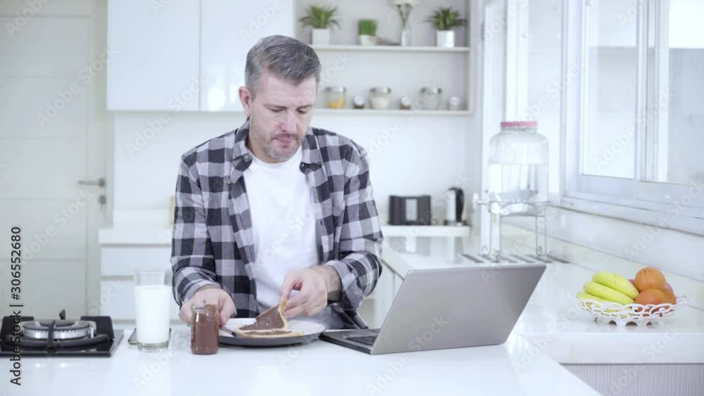 Caucasian man eating breakfast while working with a laptop in the kitchen. Shot in 4k resolution Caucasian man eating breakfast while working with a laptop in the kitchen. Shot in 4k resolution
