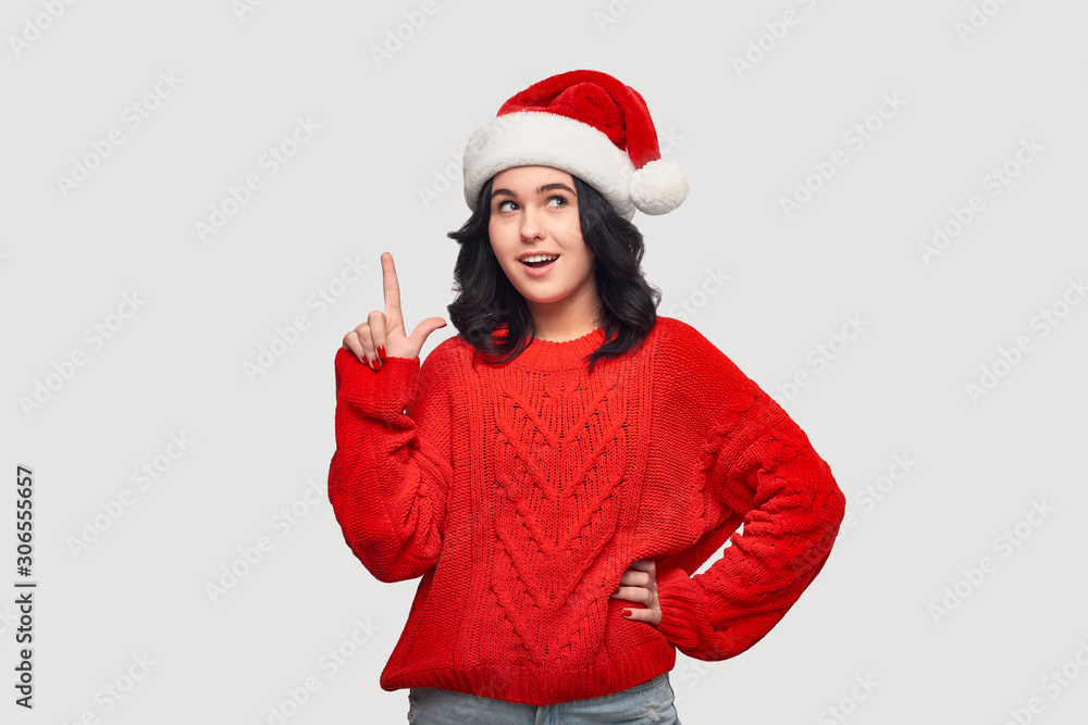 Pretty girl in a red sweater and Santa hat pointing at the copy space up over grey background Pretty girl in a red sweater and Santa hat pointing at the copy space up over grey background