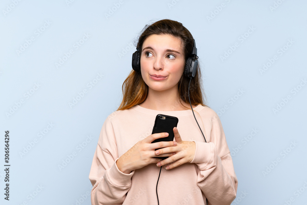 Young brunette girl over isolated blue background using the mobile with headphones and thinking Young brunette girl over isolated blue background using the mobile with headphones and thinking