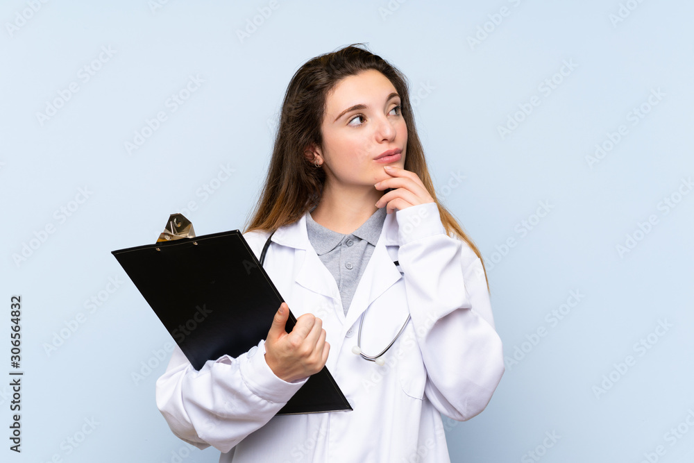 Young brunette girl over isolated blue background with doctor gown and holding a folder while thinking Young brunette girl over isolated blue background with doctor gown and holding a folder while thinking