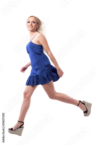 Shopping rush concept. Young stylish blonde girl in blue dress running and smiling at camera. Full body isolated on white background. 