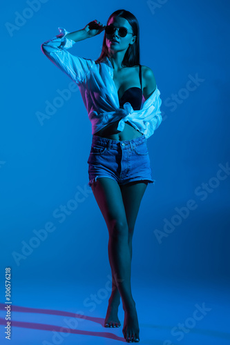 Full height of young sexy woman in shorts and white shirt, sunglasses standing on blue background