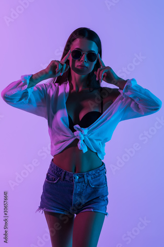 Young sexy woman in short and shirt, sunglasses standing isolated on pink light background