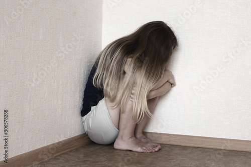 Lonely sad young blond haired girl is sitiing in the corner crying and holding her knees. Depressed woman is sitting in the corner of the room. Concept of domestic violence, abuse and harassment