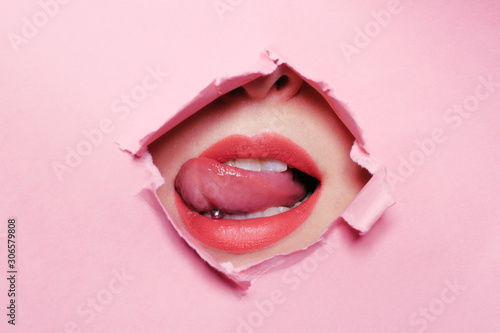 Girl bites her tongue, tongue piercing. Big beautiful lips, piercing parlor. Beautiful teeth, dentistry. Pink background, girl's face through torn cardboard. photo