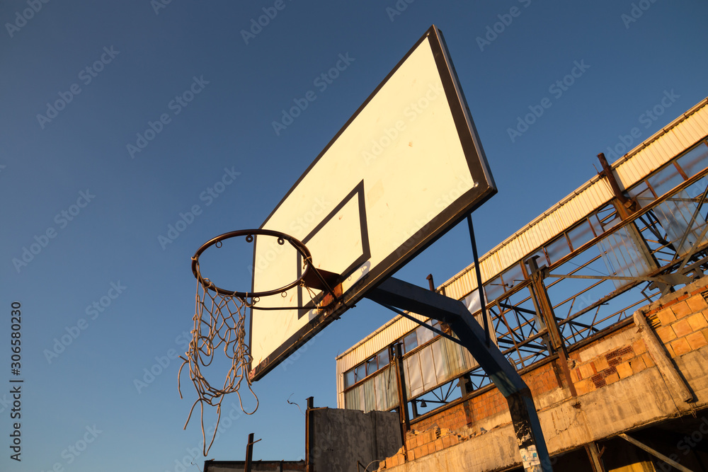 Broken basketball net dangling from a hoop attached to the white backboard. Defunct playground in recreational area, next to an abandoned and demolished industrial hall. Sunny day and clear blue sky. Broken basketball net dangling from a hoop attached to the white backboard. Defunct playground in recreational area, next to an abandoned and demolished industrial hall. Sunny day and clear blue sky.