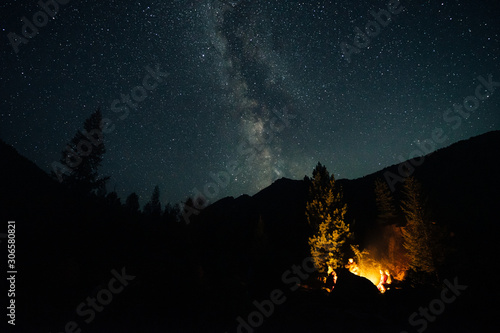 Friends sit around the fire at night and talk. Camp on a mountain hike. Starry sky over the forest. Milky Way. A romantic evening. Active tourism.