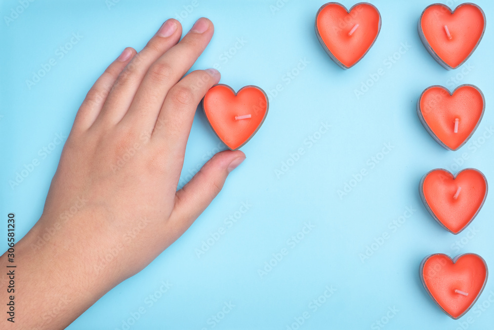 Fototapeta premium Flat lay a red candle in the form of a heart in children's hands and the remaining candles are located nearby isolated on light blue backgound. Wedding card ideas and Valentines Day