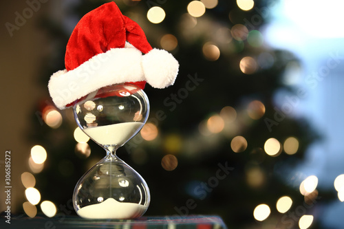 Christmas countdown. Modern Hourglass with space for text -- sand trickling through the bulbs of a crystal sand glass. Christmas tree & lights as background. Holiday-themed image.