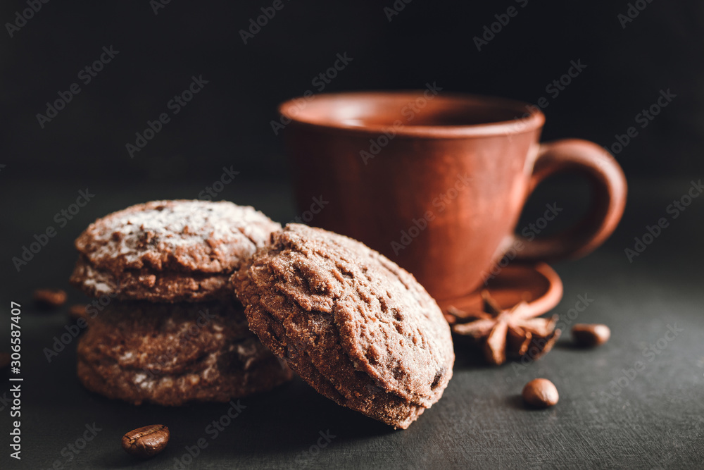 Chocolate cookies with powdered sugar and brown cup with hot drink, coffee or tea on dark background Chocolate cookies with powdered sugar and brown cup with hot drink, coffee or tea on dark background