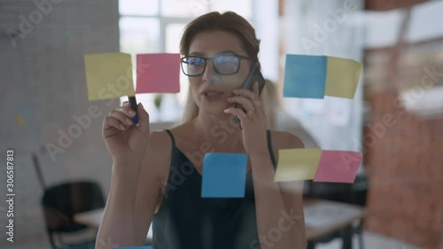 portrait of a successful girl medium shot, serious business woman talking on the phone and taking notes on stickers. modern office with glass wall 4K