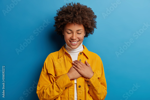 Tender feminine woman keeps hands on heart, has grateful look, appreciates effort, has eyes closed, wears yellow shirt, isolated over blue background, receives touching compliment, smiles pleasantly photo