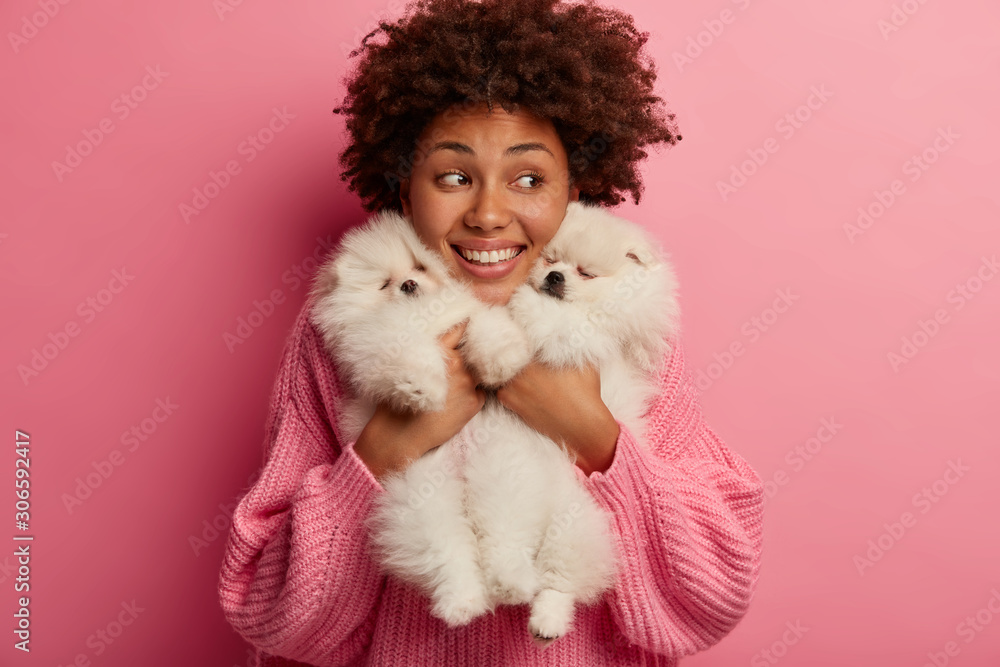 Happy Afro American woman caressing gently her dogs, holds two pets closely to face, gazes aside, tranis animals during walk, watches something with curioisity, have fun together, play around Happy Afro American woman caressing gently her dogs, holds two pets closely to face, gazes aside, tranis animals during walk, watches something with curioisity, have fun together, play around