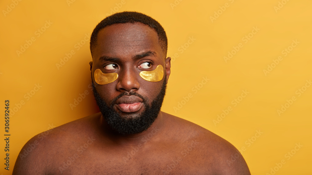 Serious Afro man has healthy dark skin, collagen patches under eyes, stands naked against yellow background, has anti aging procedure, looks thoughtfully aside. Skin care and cosmetology concept Serious Afro man has healthy dark skin, collagen patches under eyes, stands naked against yellow background, has anti aging procedure, looks thoughtfully aside. Skin care and cosmetology concept