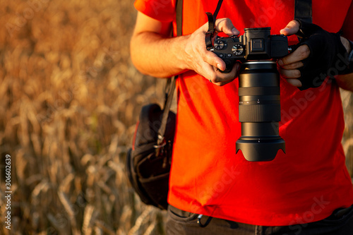 Close up man holding modern digital camera with big zoom lens while shooting in wheat field at sunset. The photographer views the captured images on the camera