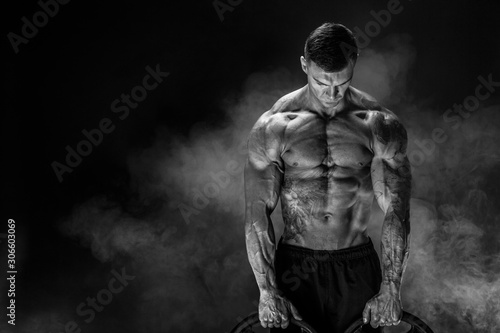 Very brawny guy bodybuilder. Bodybuilder with dumbbells in his arms on dark background with smoke.
