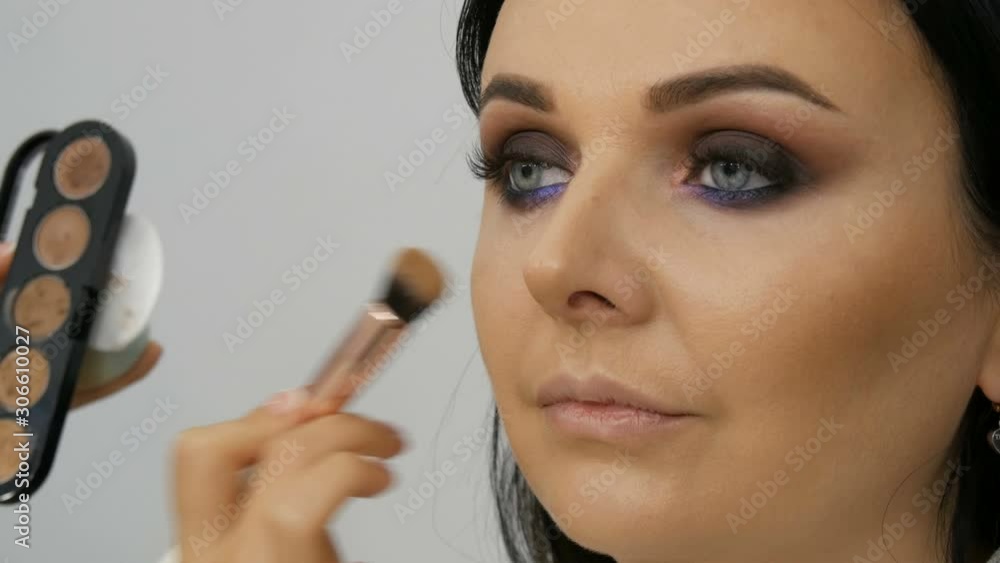 Stylist makeup artist applies foundation for face with a special brush on the face of a young beautiful woman with blue eyes