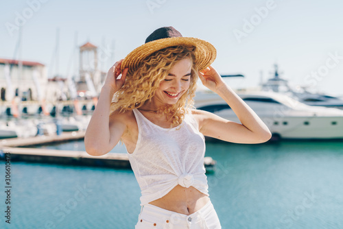 the girl is a blond angel with curly hair  resting her eyes tenderly smiling and holding her hat from the wind  behind the sea and ships
