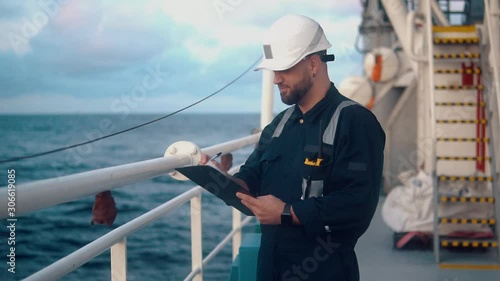 Marine chief officer or chief mate on deck of ship or vessel. He fills up ahts vessel checklist. Ship routine paperwork. He holds VHF walkie-talkie radio in hands.