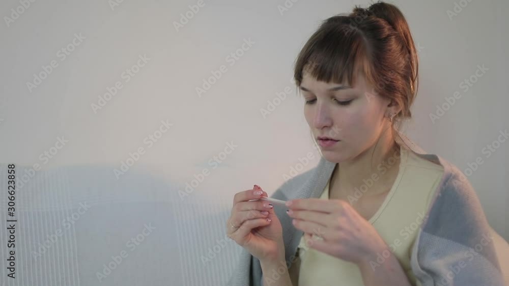 A young woman shows symptoms of a cold. She has a fever, cough and chills. A girl wraps herself in a plaid and puts on a knitted hat. A woman takes and drinks pills.