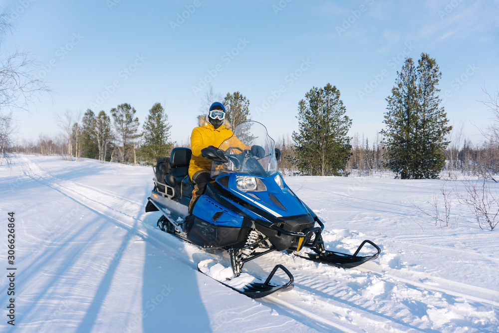 young guy in a yellow warm jacket sits on his snowmobile and rides through an empty snowy meadow surrounded by trees on a frosty winter day