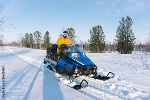 young guy in a yellow warm jacket sits on his snowmobile and rides through an empty snowy meadow surrounded by trees on a frosty winter day