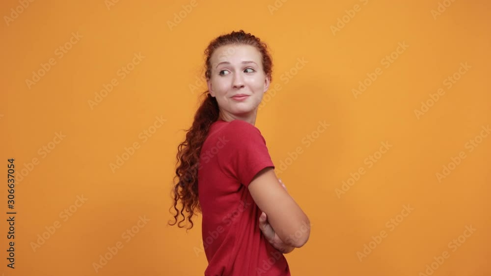 Pretty young woman in red shirt holding hands crossed looking aside isolated on orange background in studio. People sincere emotions, health concept. Pretty young woman in red shirt holding hands crossed looking aside isolated on orange background in studio. People sincere emotions, health concept.