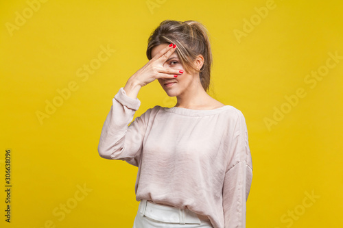 Portrait of curious beautiful young woman with fair hair in casual beige blouse standing, peeking through fingers with suspicious and prying eyes. indoor studio shot isolated on yellow background