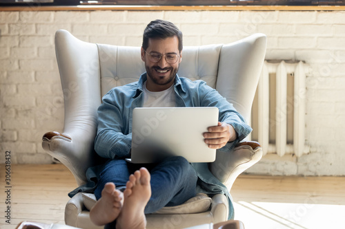 Smiling relaxed young man sit on armchair using laptop laughing photo