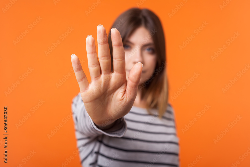 Portrait of young brunette woman in long sleeve striped shirt standing with serious look and showing stop hand gesture at camera, refusal or prohibition. indoor shot isolated on orange background Portrait of young brunette woman in long sleeve striped shirt standing with serious look and showing stop hand gesture at camera, refusal or prohibition. indoor shot isolated on orange background