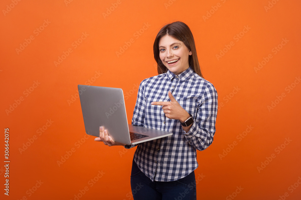 Portrait of smart positive woman with brown hair in checkered casual shirt and denim standing pointing at laptop in her hands, smiling at camera. indoor studio shot isolated on orange background Portrait of smart positive woman with brown hair in checkered casual shirt and denim standing pointing at laptop in her hands, smiling at camera. indoor studio shot isolated on orange background