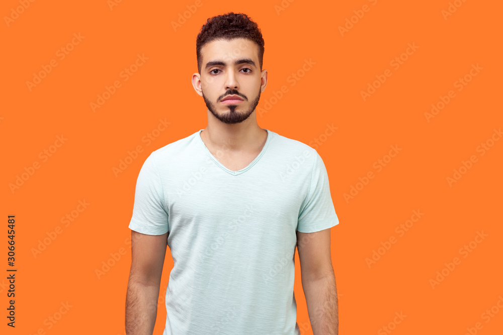 Portrait of serious handsome brunette man with beard in casual white t-shirt standing with hands down and looking focused and confident at camera. indoor studio shot isolated on orange background Portrait of serious handsome brunette man with beard in casual white t-shirt standing with hands down and looking focused and confident at camera. indoor studio shot isolated on orange background