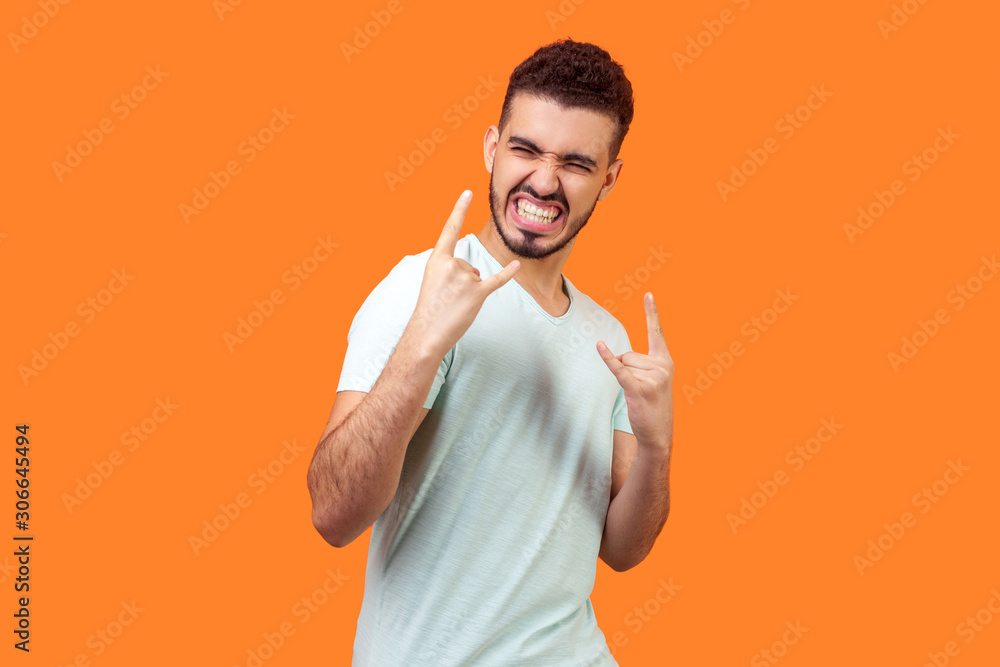 Portrait of excited crazy brunette man with beard in casual white t-shirt showing devil horns gesture with aggressive grimace, rock n roll punk sign. indoor studio shot isolated on orange background Portrait of excited crazy brunette man with beard in casual white t-shirt showing devil horns gesture with aggressive grimace, rock n roll punk sign. indoor studio shot isolated on orange background