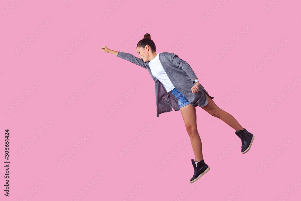 Superman. Portrait of cheerful ambitious brunette teenage girl with bun hairstyle in casual clothes flying with one raised hand, feeling super power. indoor studio shot isolated on pink background