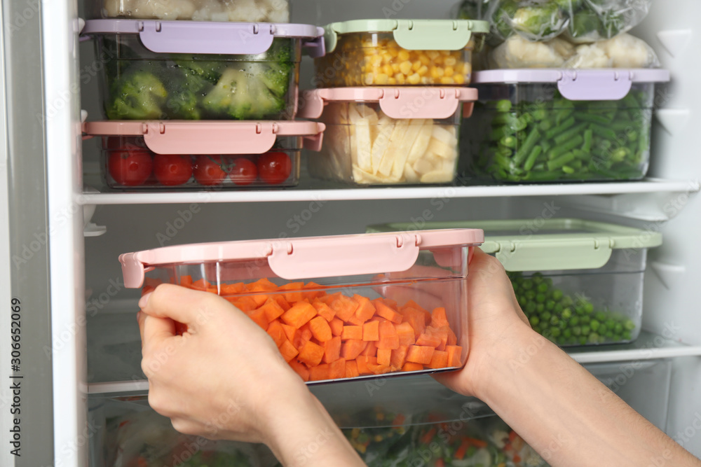 Woman putting container with carrot in refrigerator with frozen vegetables, closeup