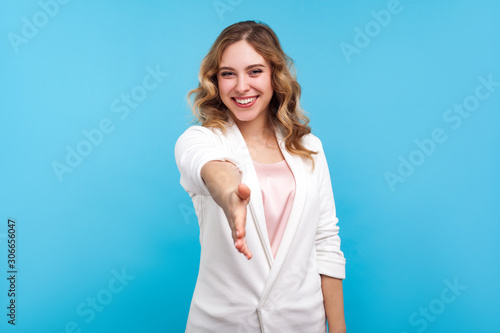 Welcome! Portrait of friendly hospitable cheerful woman with wavy hair in white jacket giving hand to handshake, hostess greeting guests, looking sociable positive. indoor studio shot, blue background