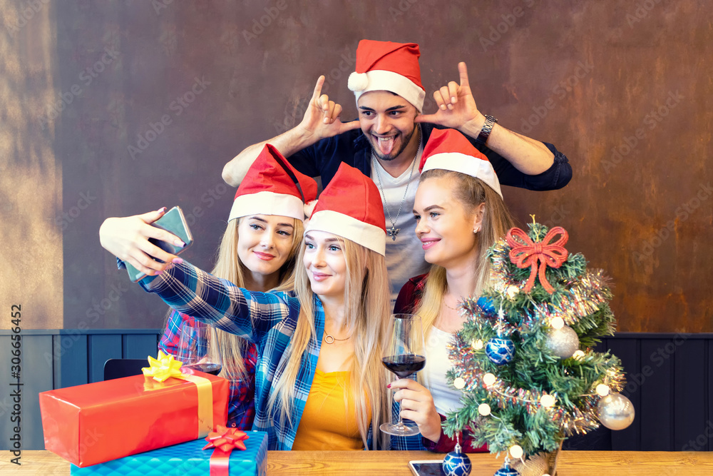 Obraz premium Happy young friends wearing Santa claus hat enjoying Christmas party taking selfie with funny faces at home sharing it on social media
