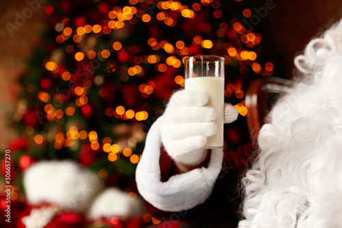 Santa Claus holding glass of milk against blurred Christmas lights, closeup photo