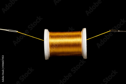 A spool of gold thread suspended on a field of black on a thread between 2 needle eyes. Plenty of space for copy.