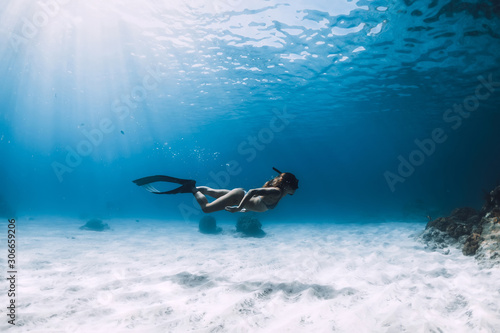 Freediver girl with fins glides over sandy bottom with fishes in blue ocean