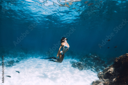 Freediver girl with fins glides over sandy bottom with fishes in blue ocean
